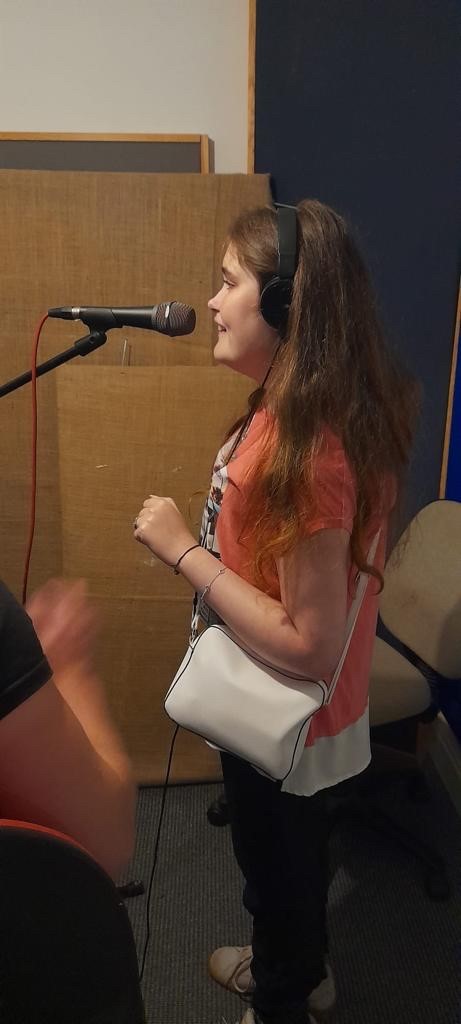 Grace is stood in a studio singing happily into a microphone. She has long brown hair and wears a peach t-shirt and holds a white handbag.