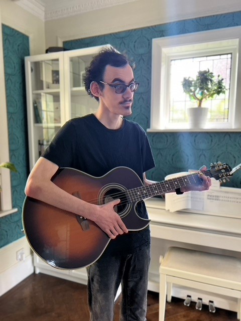 Ben is standing in a living room holding an acoustic guitar ready to play. He has dark hair and wears a black t-shirt.