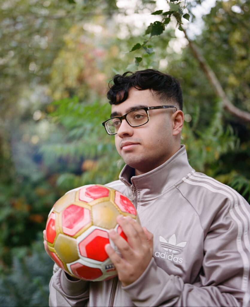 Bilaal stands outside holding a football which is red and gold. He wears a grey Adidas tracksuit top. 