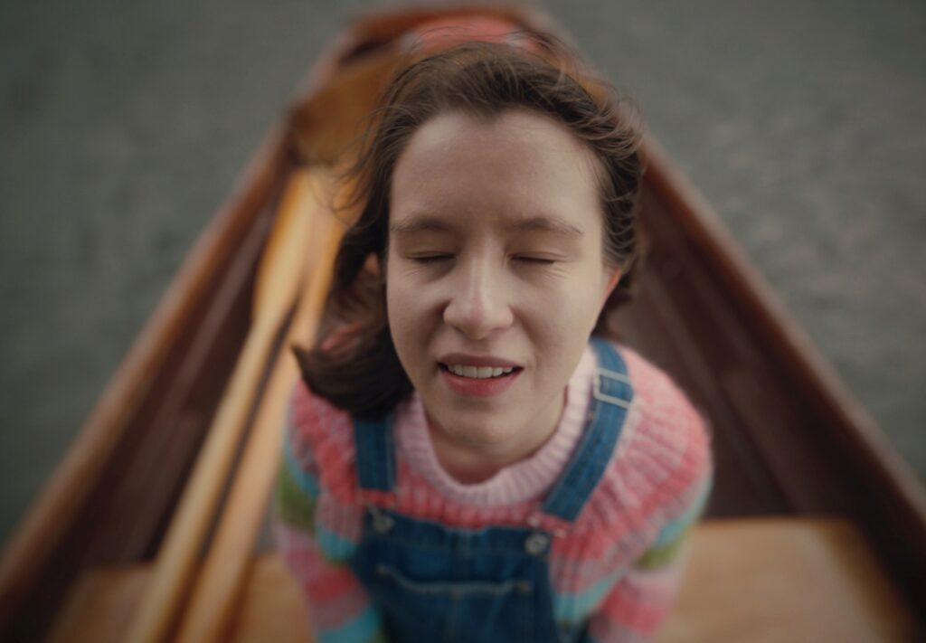 Ellie is facing towards the sky smiling with her eyes closed and wind blowing through her hair. She is wearing a knitted jumper and dungarees and standing in a wooden boat with oars. It is a very atmospheric image with a movie like quality.