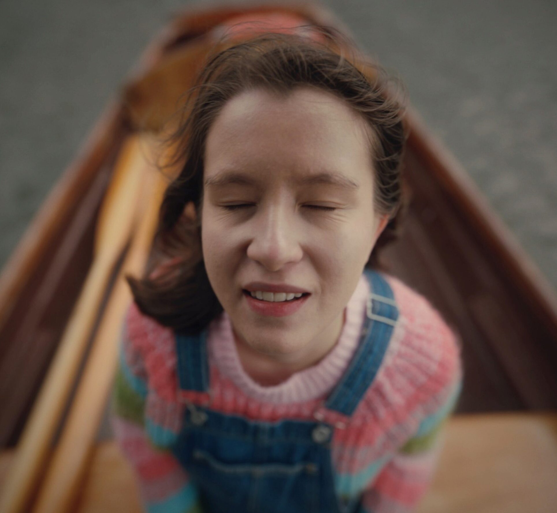 Ellie is facing towards the sky smiling with her eyes closed and wind blowing through her hair. She is wearing a knitted jumper and dungarees and standing in a wooden boat with oars. It is a very atmospheric image with a movie like quality.