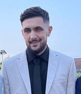 Kieran is standing outside dressed smartly for an occasion. He is wearing a black shirt and a beige dinner jacket. 