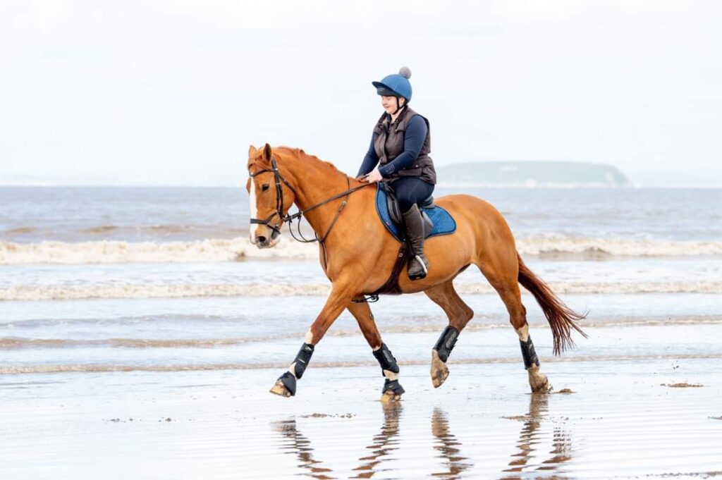 Lucy is riding a horse on a beach with the sea behind. The horse is a tan colour and has a white stripe on their nose. Lucy wears riding gear and a horse riding hat with a bobble on top.