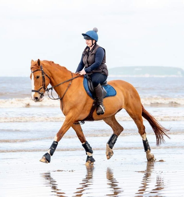 Lucy is riding a horse on a beach with the sea behind. The horse is a tan colour and has a white stripe on their nose. Lucy wears riding gear and a horse riding hat with a bobble on top.