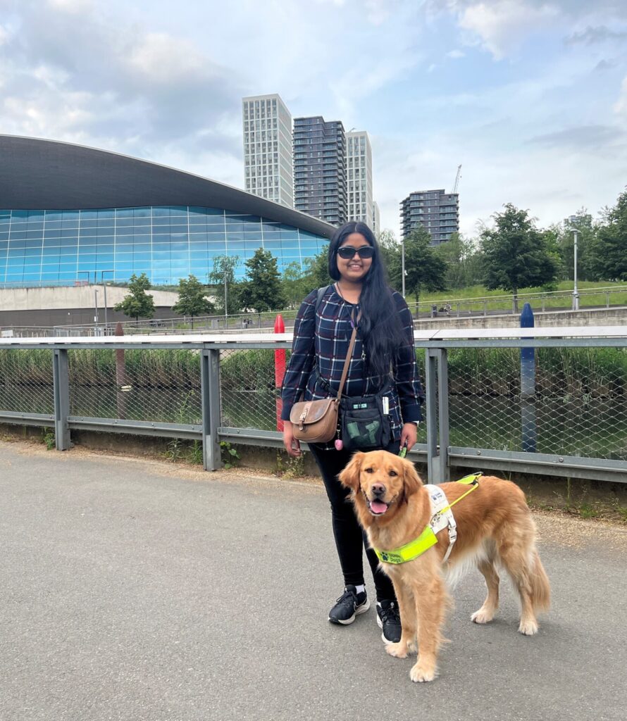 Me and my Guide Dog Bertie. Bertie is a golden retriever he is wearing his Guide Dogs harness standing by my side smiling at the camera. I am stood next to him I have long dark hair, brown skin, wearing sunglasses, A full sleeves dark top and a pair of black geans. This photo was taken outside there is a small canal in the background and a building towards the back on the left.