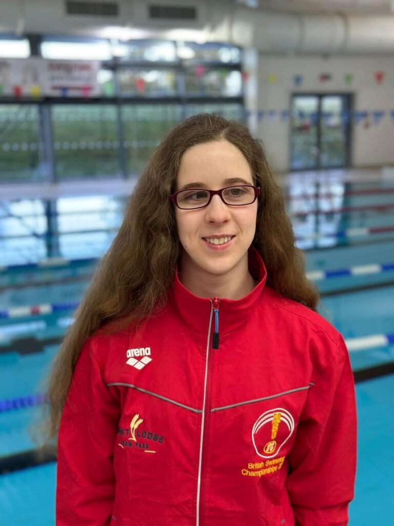 Scarlett is smiling standing in front of an indoor swimming pool. She has long brown hair and wears glasses and a red zip-up British Swimming Championship tracksuit top.