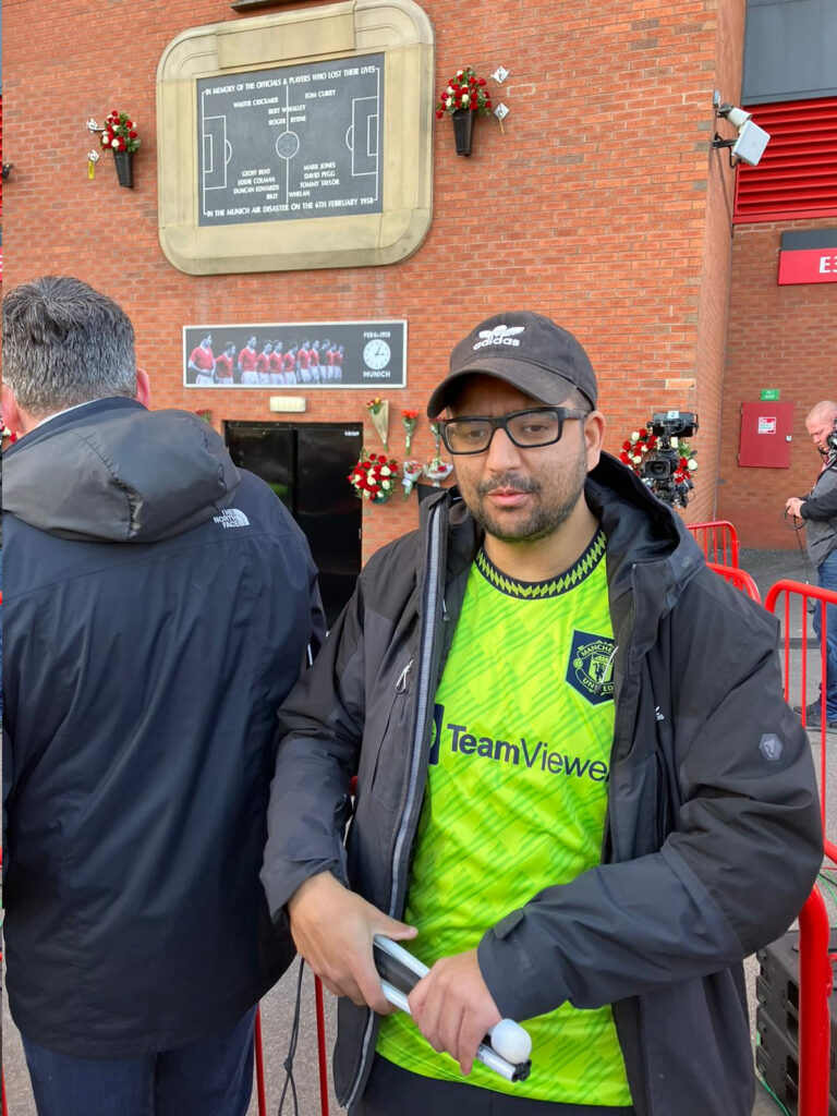 Abu is wearing a black cap and a green Manchester United shirt and a black jacket. He is standing outside the grounds and is holding a folded up white cane.