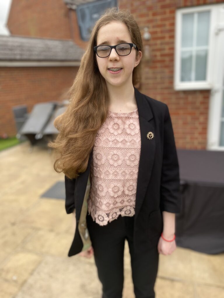 Eliza is standing on her patio smiling, she is smartly dressed wearing a light pink laced top and a black blazer. She has long brown hair and wears glasses.