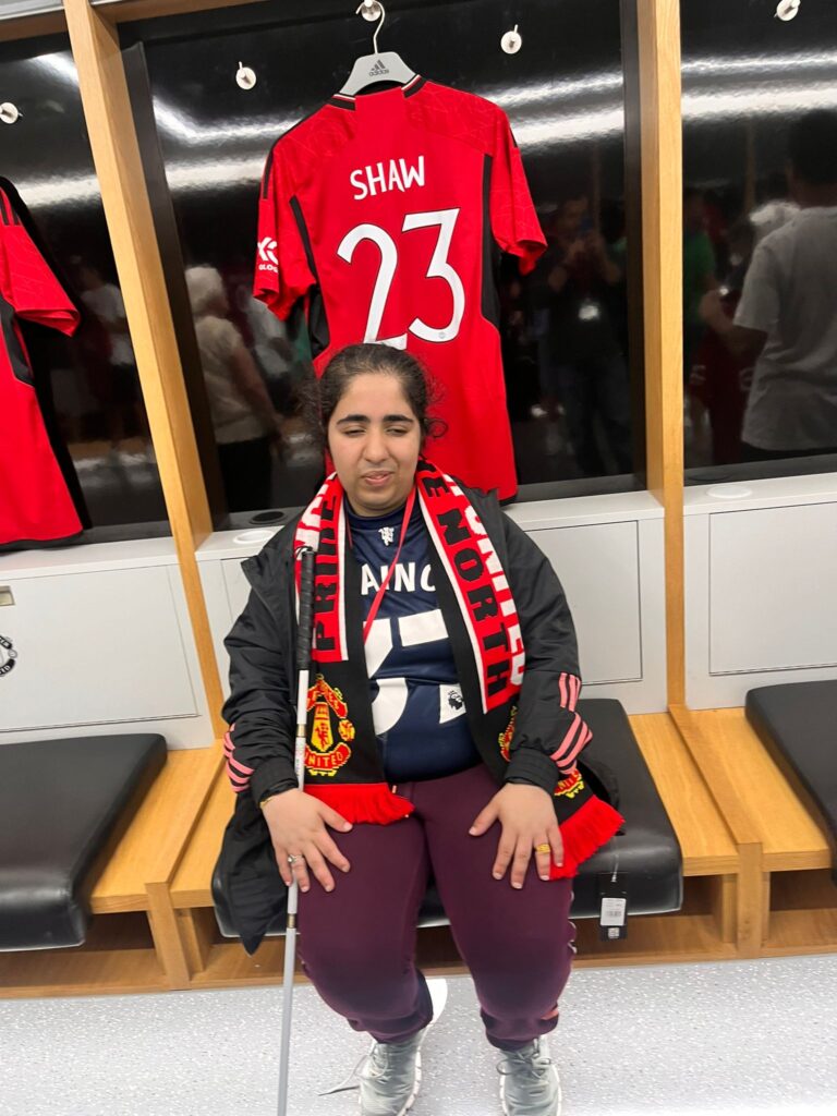  This pic was of me at Man United stadium as I am a united fan. Ikra sits on a bench with a Man United shirt hanging behind her - the shirt has the number 23 and the name SHAW. Ikra is wearing a Man United scarf and is holding her white cane. 