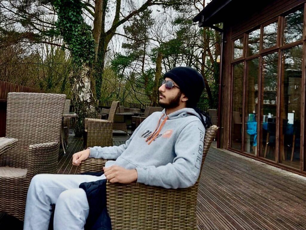 Kiranjeet is relaxing in a wicker chair out on some decking. He is wearing a grey Nike tracksuit and a black beanie and sunglasses, looking off into the distance.