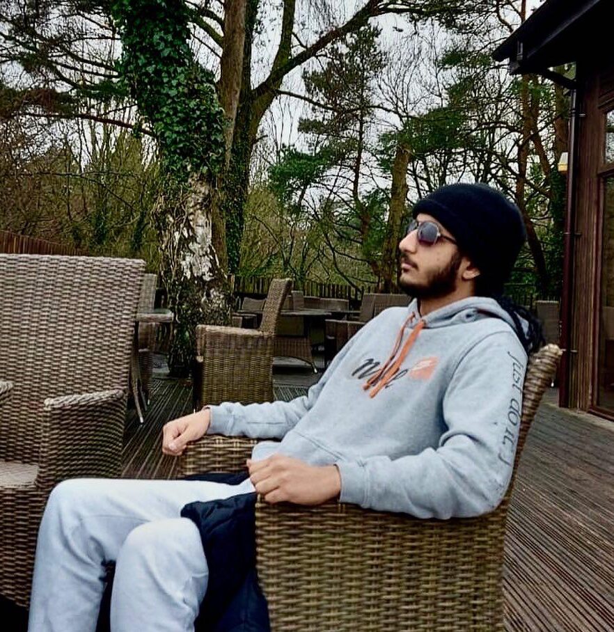 Kiranjeet is relaxing in a wicker chair out on some decking. He is wearing a grey Nike tracksuit and a black beanie and sunglasses, looking off into the distance.