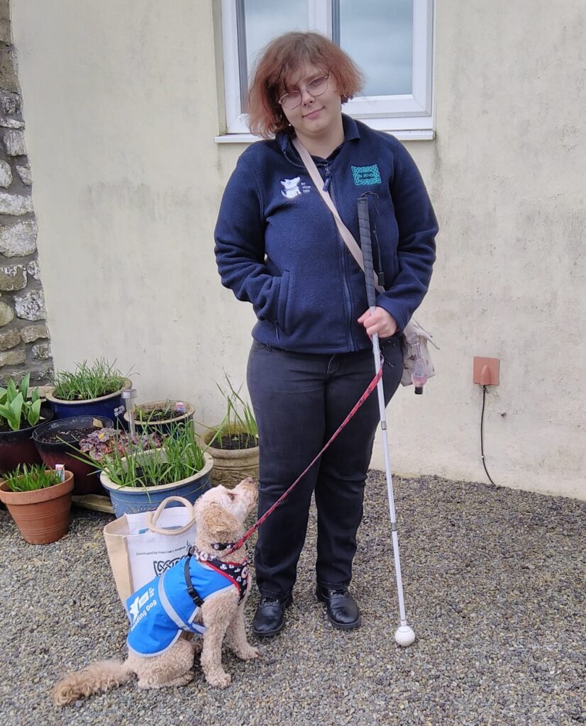 Rhiana stands outside holding her white cane. She is wearing a navy hoodie and has short brown hair. Next to her Rhiana's cavapoo looks up at her. He is wearing a blue coat.