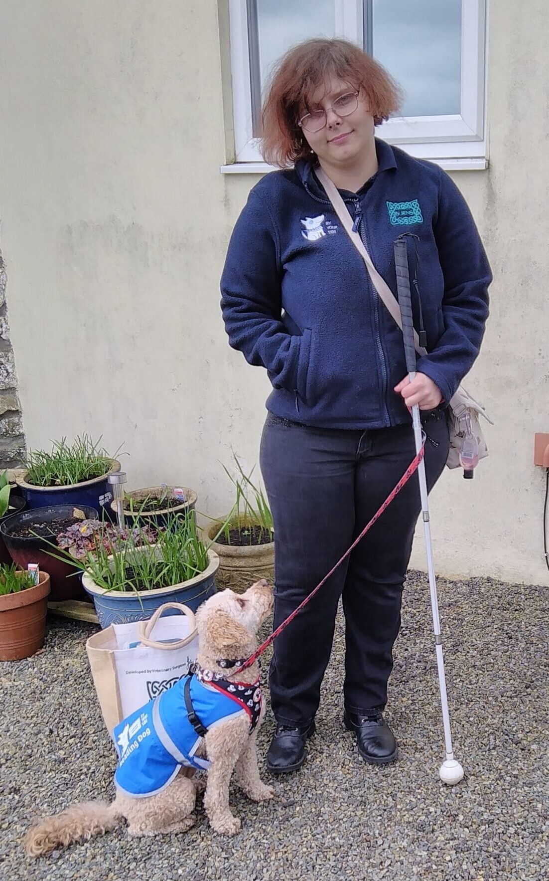 Rhiana stands outside holding her white cane. She is wearing a navy hoodie and has short brown hair. Next to her Rhiana's cavapoo looks up at her. He is wearing a blue coat.