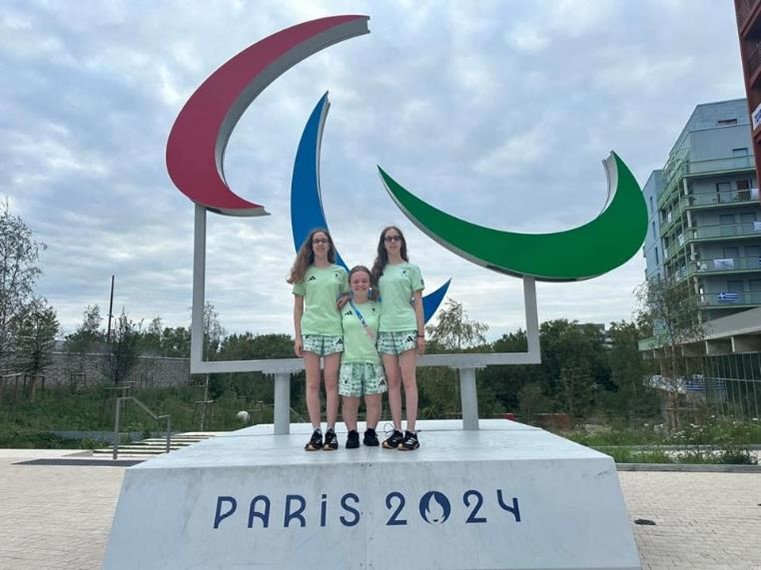 Eliza, Scarlett and Maisie are standing on the base of the statue which reads 'Paris 2024'. Behind them is the “Agitos” symbol which is shaped like three moon crescent shapes which are red, blue and green.