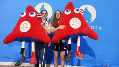 Eliza and Scarlett holding the Paralympic “Phryges” mascot. The mascots are giant soft toys almost as tall as they are and they are holding one each. They are red triangles with faces and dangly legs.