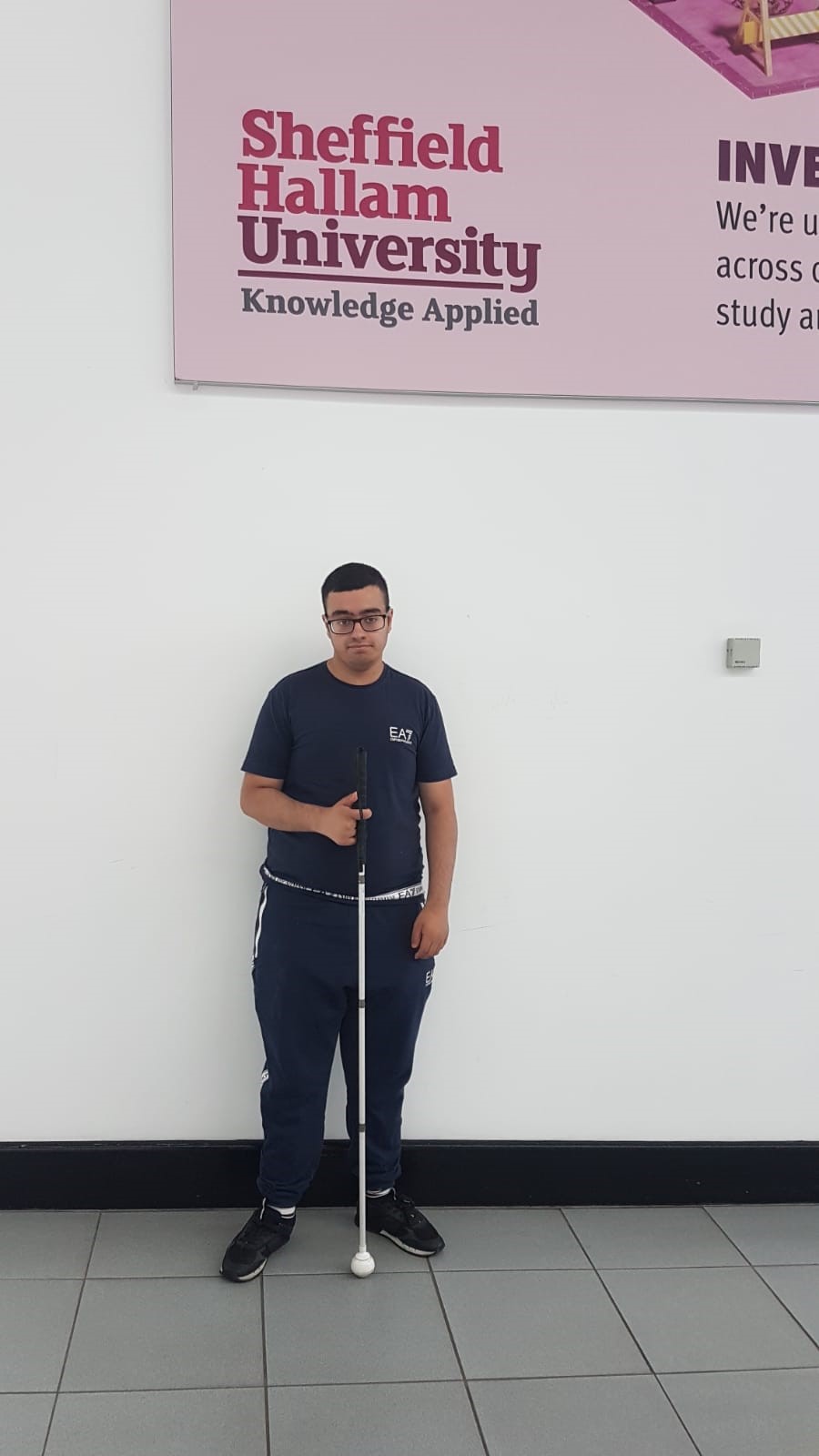 Bilaal is holding his white cane and standing in front of a white wall with a large Sheffield Hallam University sign on it