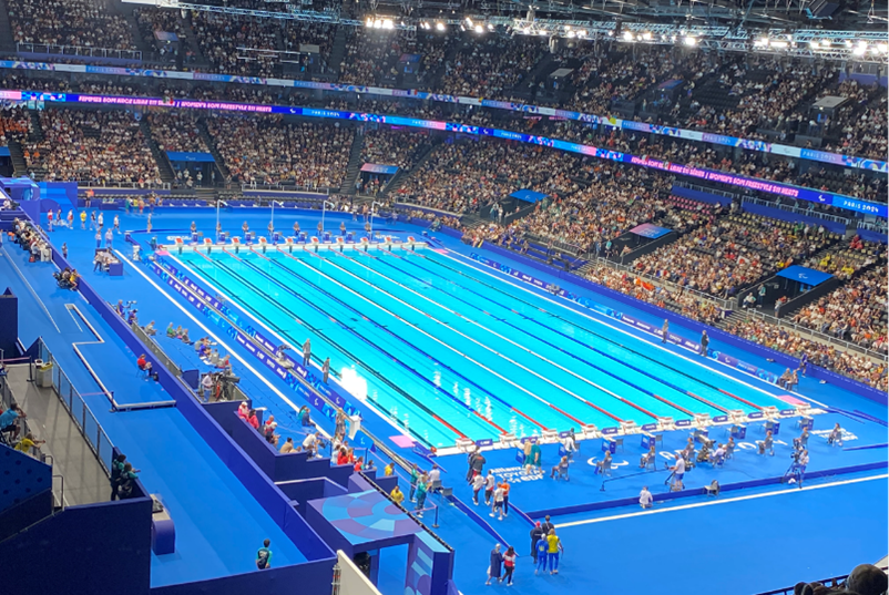 Looking down on the Olympic swimming pool inside the arena in Paris. The stadium is full of thousands of people. The swimmers are walking out to the podium.