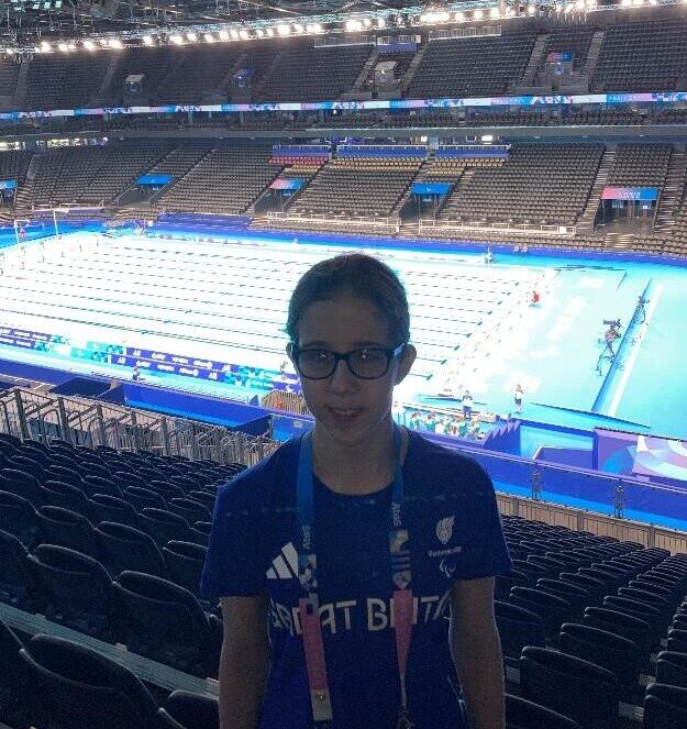 Eliza is wearing a blue Great Britain t-shirt and lanyard. She is standing at the stop of the tiered seating which is empty. Behind and below is the swimming pool.