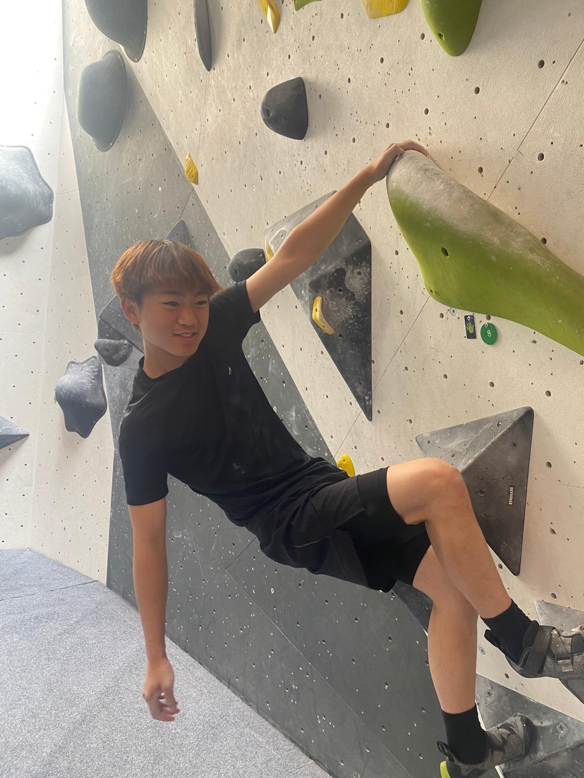 Johnathan free climbing at an indoor climbing wall. One hand holds the grip and the other dangles down as he smiles to camera.
