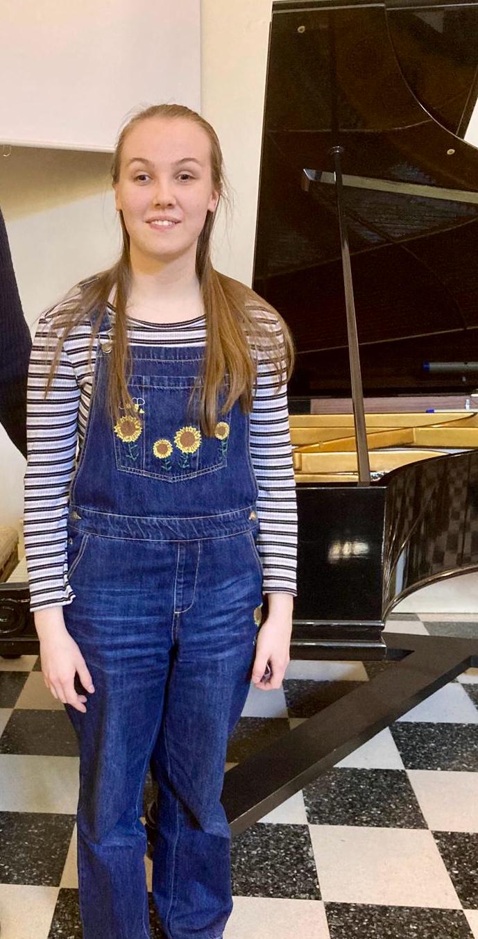 Lydia stands smiling in front a grand piano. She wears denim dungarees sunflowers embroidered on.