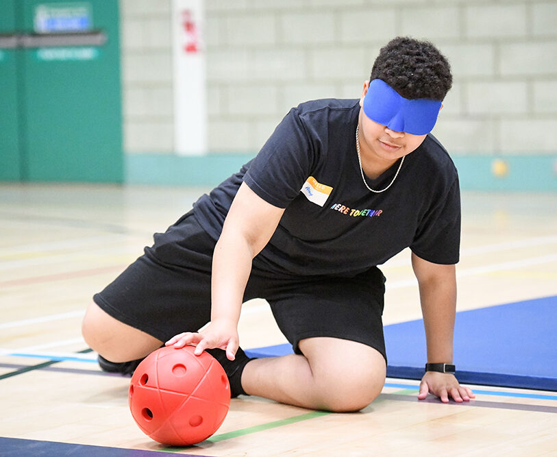Alex playing goalball blindfolded. He is in an sports hall, down on the floor with one hand on the ball.