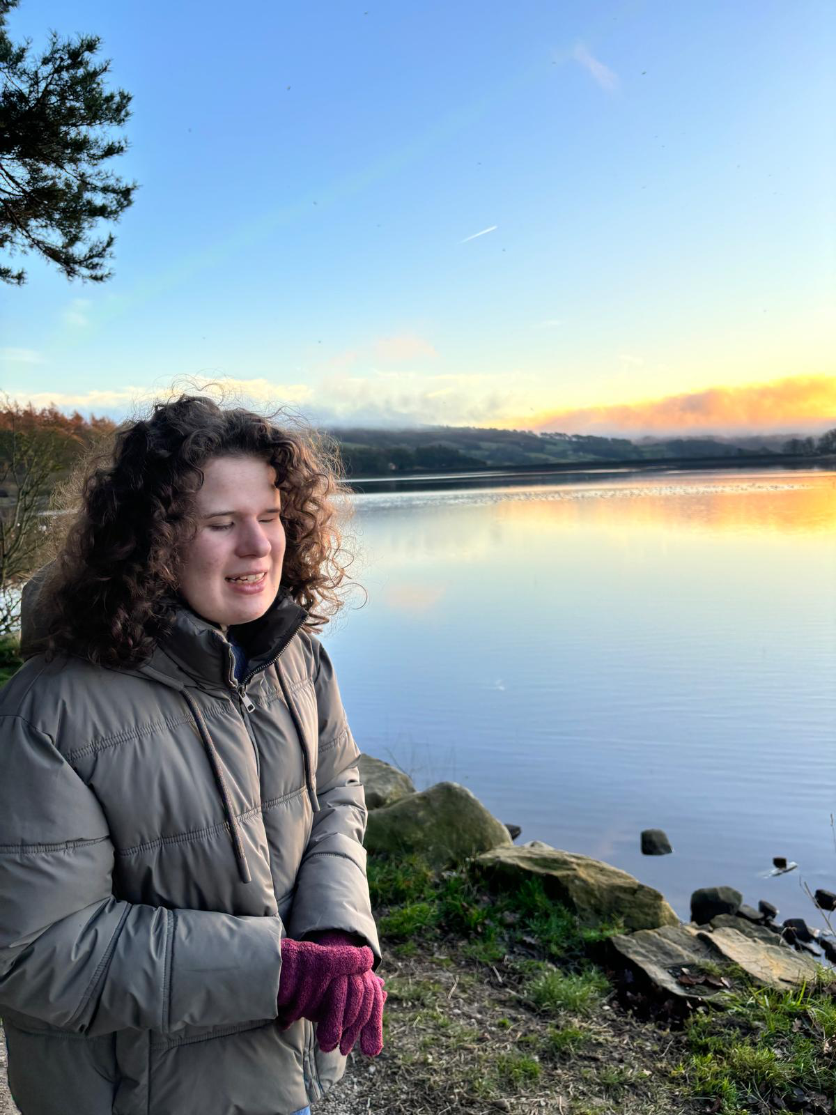 Lydia wears winter clothes and smiles looking over a beautiful still lake with a sunset in the background