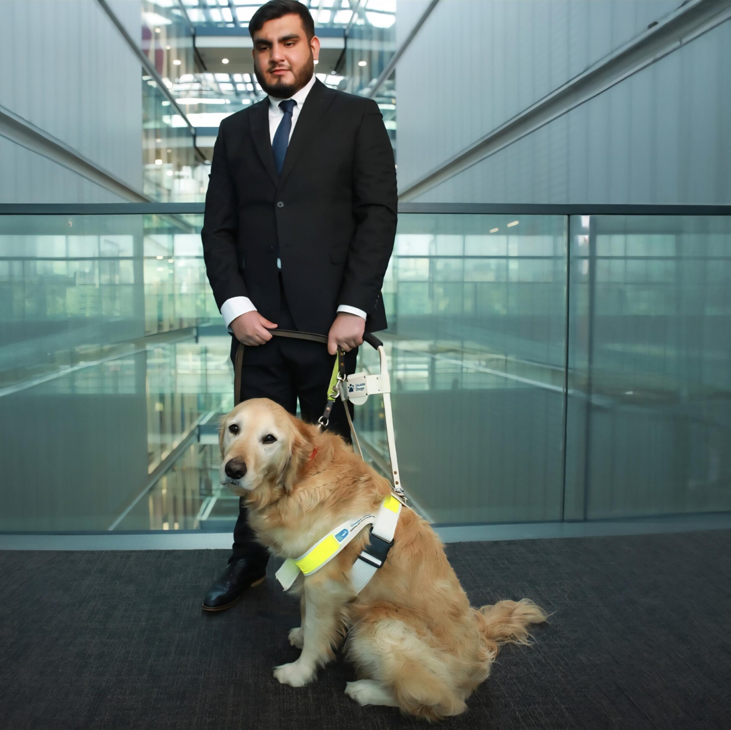 Ravi wears a smart suit and stands on a mezzanine layer of a modern office with his golden retriever guide dog.