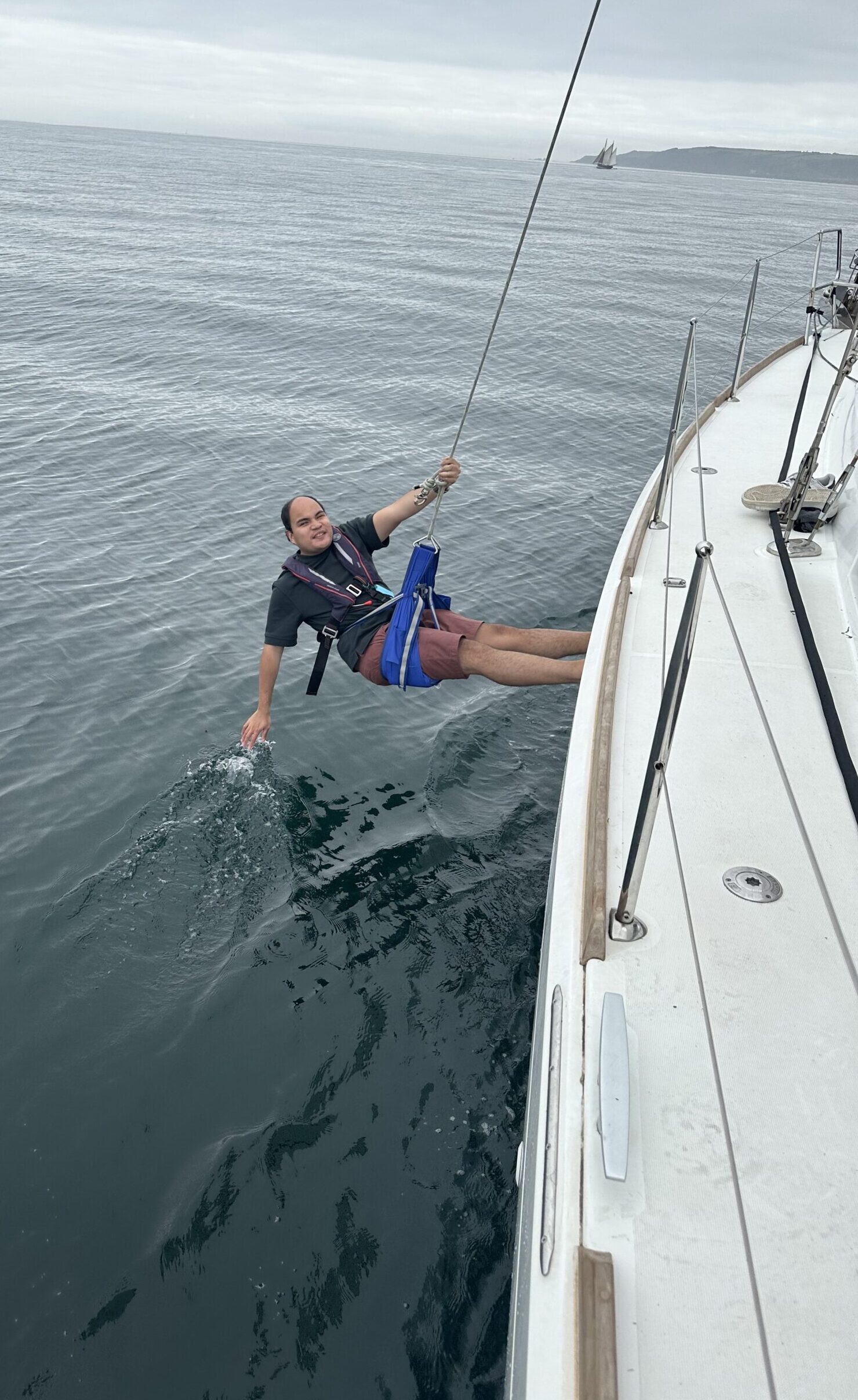 Robbin hangs from the side of a sailing boat. He holds the rope with one hand and trails the other across the water.