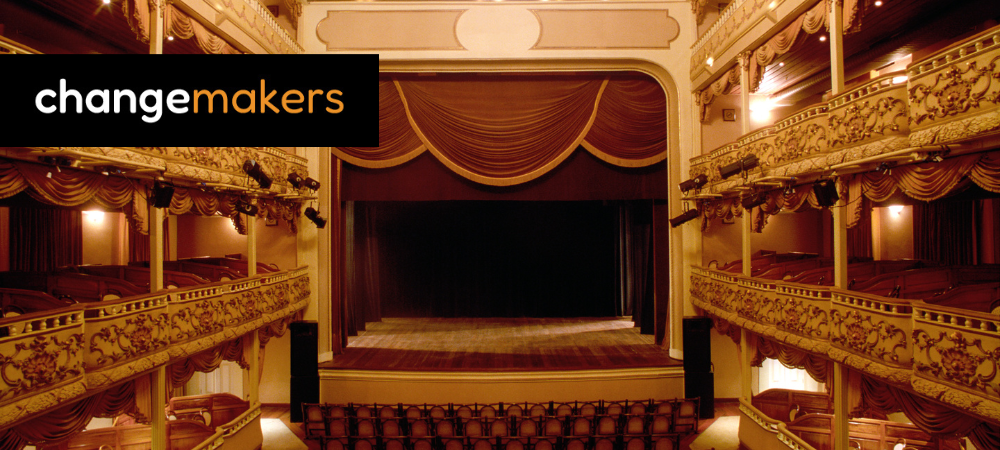 An empty theatre with red velvet seats and a red curtain covering a proscenium arch. The changemakers logo is top left.