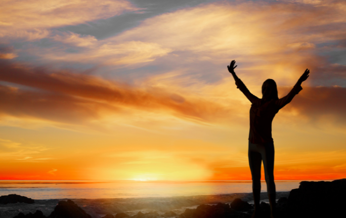 Woman stretching up to the sky joyously, backlit by a sunset.