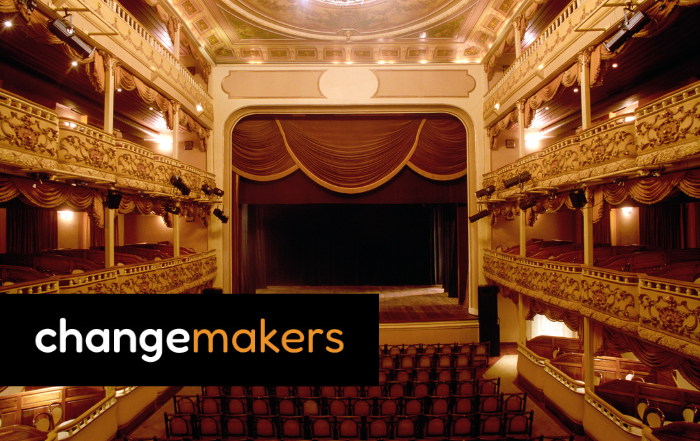 An empty theatre with red velvet seats and curtain covering a proscenium arch with gold details.